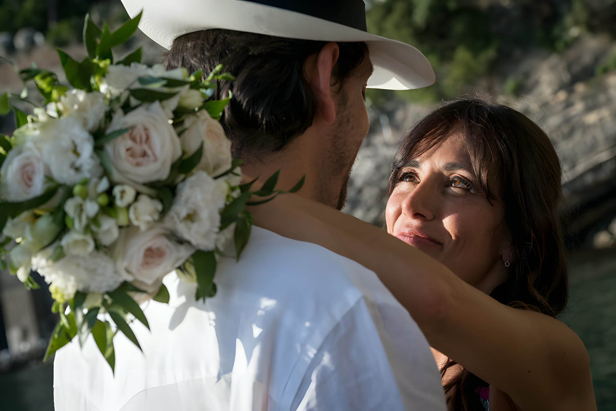 Ritratto ravvicinato: la sposa con un cappello a tesa larga e rose bianche abbraccia lo sposo